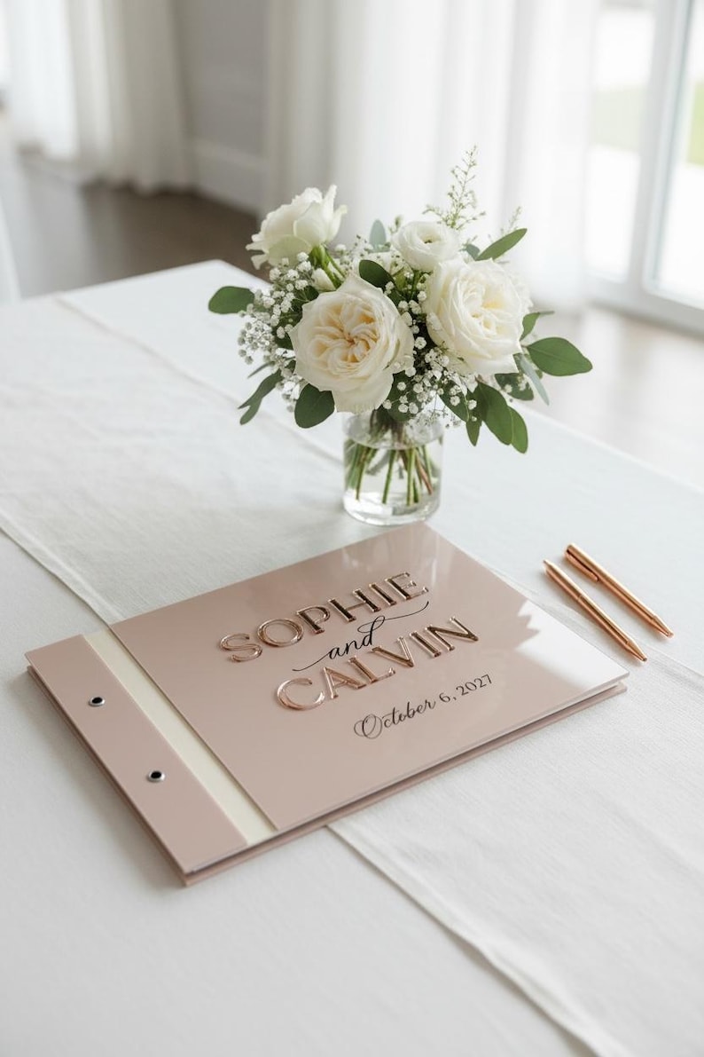 May include: A blush pink guest book with a glossy finish featuring the embossed text "SOPHIE and CALVIN" and "October 6, 2027" in a metallic rose gold font. The book has a side binding with two silver metal rivets and two rose gold pens.