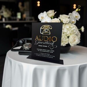Puede incluir: Un letrero negro con letras doradas y un gr&aacute;fico de tel&eacute;fono vintage que dice "Audio Guestbook". El letrero est&aacute; sobre un mantel blanco, con un tel&eacute;fono negro y un ramo de flores blancas en el fondo.