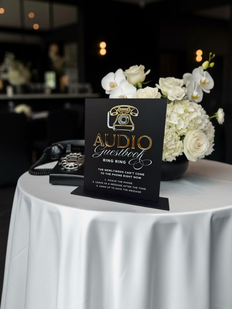 Puede incluir: Un letrero negro con letras doradas y un gr&aacute;fico de tel&eacute;fono vintage que dice "Audio Guestbook". El letrero est&aacute; sobre un mantel blanco, con un tel&eacute;fono negro y un ramo de flores blancas en el fondo.