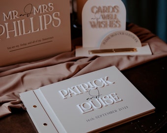 May include: A white wedding guest book with the names Patrick and Louise and the date 16th September 2023 embossed on the front cover. The book is closed and sitting on a brown wooden table.