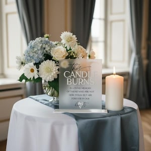 K&ouml;nnte beinhalten: Ein mattes Acryl-Gedenkschild mit schwarzem Text This CANDLE BURNS IN LOVING MEMORY OF THOSE WHO ARE NOT HERE TODAY BUT ARE FOREVER IN OUR hearts, neben einer wei&szlig;en Stumpenkerze und einem Blumenstrau&szlig; aus wei&szlig;en und blauen Bl&uuml;ten.