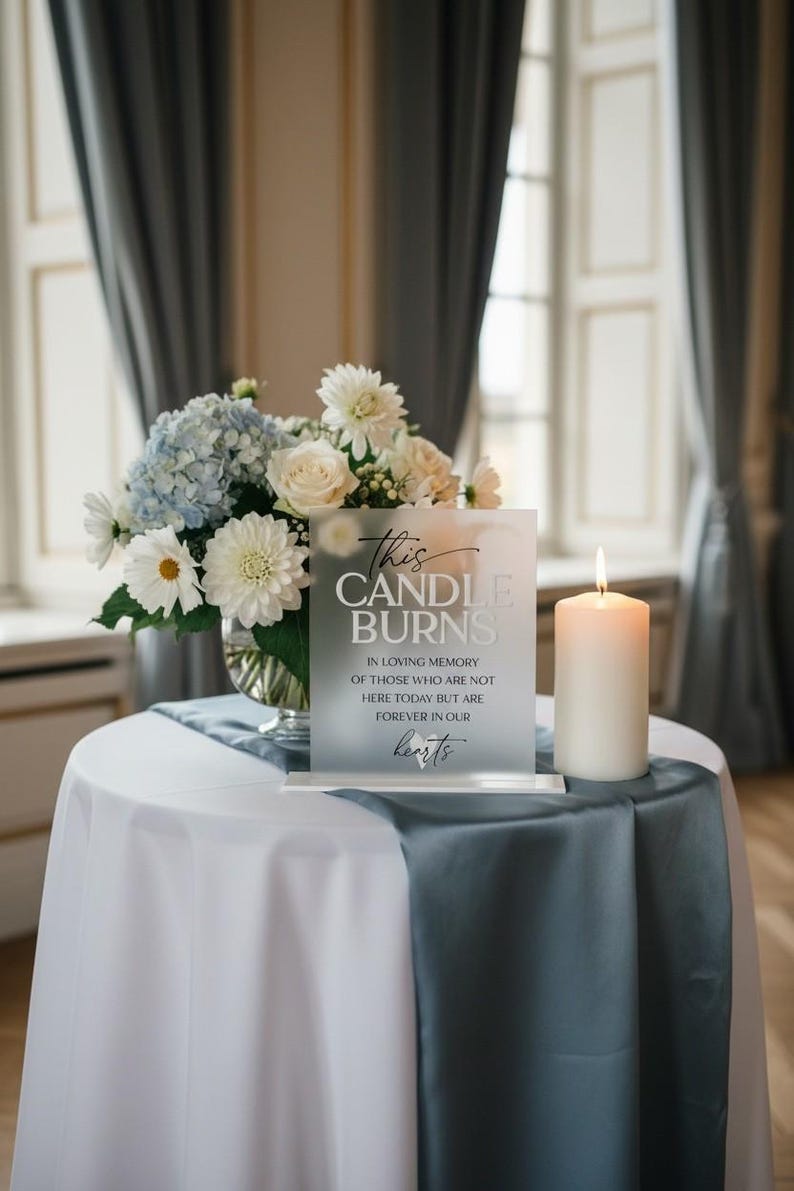 K&ouml;nnte beinhalten: Ein mattes Acryl-Gedenkschild mit schwarzem Text This CANDLE BURNS IN LOVING MEMORY OF THOSE WHO ARE NOT HERE TODAY BUT ARE FOREVER IN OUR hearts, neben einer wei&szlig;en Stumpenkerze und einem Blumenstrau&szlig; aus wei&szlig;en und blauen Bl&uuml;ten.