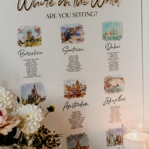 May include: A white wedding seating chart sign with gold 3D lettering reading Where in the World ARE YOU SITTING? It features watercolor illustrations of global landmarks like Barcelona, Santorini, Dubai, Amsterdam, Zakynthos, and Paris with guest lists.