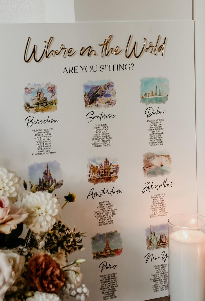 May include: A white wedding seating chart sign with gold 3D lettering reading Where in the World ARE YOU SITTING? It features watercolor illustrations of global landmarks like Barcelona, Santorini, Dubai, Amsterdam, Zakynthos, and Paris with guest lists.