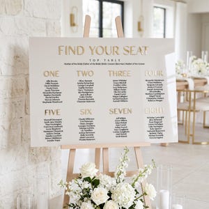 May include: A rectangular wedding seating chart sign with a white background and gold lettering. The sign features the text FIND YOUR SEAT at the top, followed by eight numbered tables with lists of names printed in black ink below each gold number.