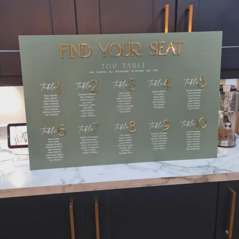 May include: A sage green wedding seating chart with gold lettering. The sign reads "FIND YOUR SEAT" and lists table numbers and guest names. The table numbers are in a gold, cursive font.