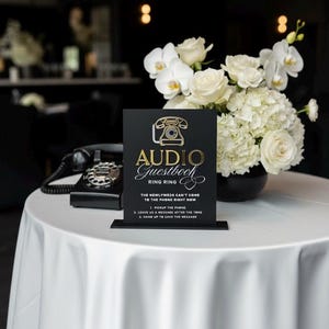 Puede incluir: Un letrero negro con letras doradas y un gr&aacute;fico de tel&eacute;fono vintage que dice "Audio Guestbook". Un tel&eacute;fono vintage negro est&aacute; sobre una mesa con un mantel blanco, junto a un arreglo floral de rosas blancas y orqu&iacute;deas.