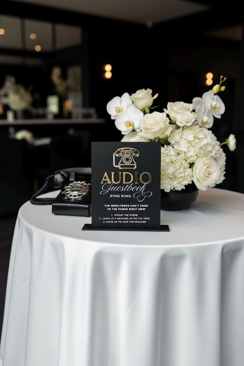 Puede incluir: Un letrero negro con letras doradas y un gr&aacute;fico de tel&eacute;fono vintage que dice "Audio Guestbook". Un tel&eacute;fono vintage negro est&aacute; sobre una mesa con un mantel blanco, junto a un arreglo floral de rosas blancas y orqu&iacute;deas.