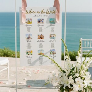 May include: A white rectangular wedding seating chart sign suspended by two dusty pink ribbons from a thin white metal frame. The sign features the gold text Where in the World ARE YOU SITTING? and nine watercolor illustrations of global locations.