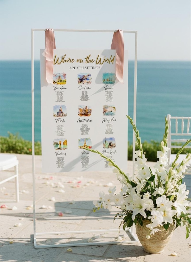 May include: A white rectangular wedding seating chart sign suspended by two dusty pink ribbons from a thin white metal frame. The sign features the gold text Where in the World ARE YOU SITTING? and nine watercolor illustrations of global locations.