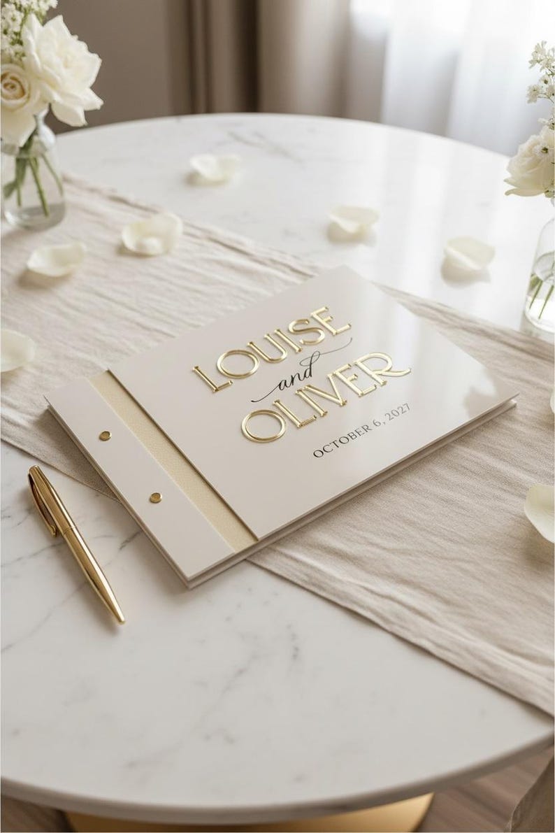 May include: A cream-colored guest book with gold embossed lettering that reads "LOUISE and OLIVER" and "OCTOBER 6, 2027". The book features a side binding with two gold metal rivets and is accompanied by a matching gold pen resting beside it.