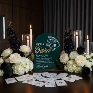 May include: A dark green arched acrylic sign with gold text reading PICK A CARD ANY CARD, PLEASE WRITE A MESSAGE AND SIGN YOUR NAME SO WE CAN REMEMBER THIS DAY WHENEVER WE PLAY, and Thank you, surrounded by white flowers and playing cards.
