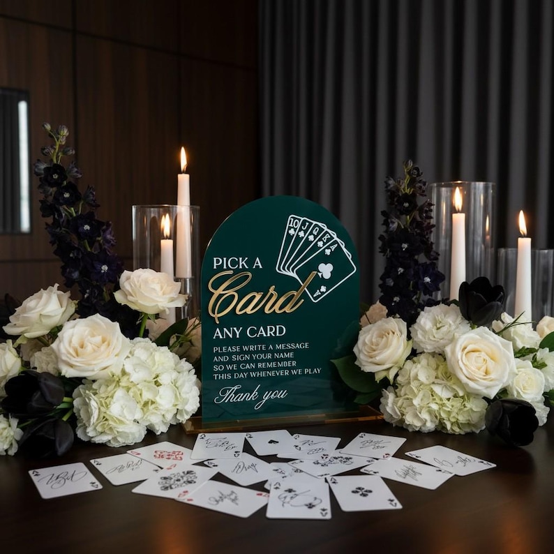 May include: A dark green arched acrylic sign with gold text reading PICK A CARD ANY CARD, PLEASE WRITE A MESSAGE AND SIGN YOUR NAME SO WE CAN REMEMBER THIS DAY WHENEVER WE PLAY, and Thank you, surrounded by white flowers and playing cards.