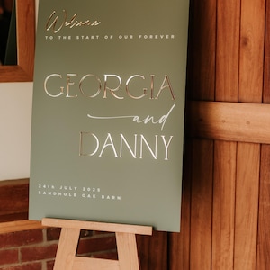 May include: A sage green wedding sign on a wooden easel. The sign features the names "GEORGIA and DANNY" in elegant lettering, along with the date "24th JULY 2025" and the venue "SANDHOLE OAK BARN". The words "Welcome to the start of our forever" are also displayed.