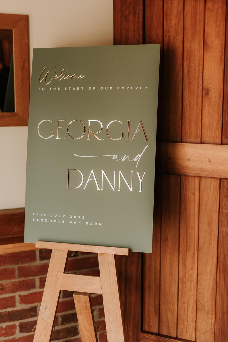 May include: A sage green wedding sign on a wooden easel. The sign features the names "GEORGIA and DANNY" in elegant lettering, along with the date "24th JULY 2025" and the venue "SANDHOLE OAK BARN". The words "Welcome to the start of our forever" are also displayed.