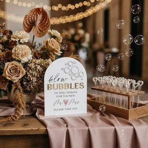 May include: A white arched sign with black text reading blow BUBBLES OF WELL WISHES FOR THE NEW Mr + Mrs PLEASE TAKE ONE, accompanied by a wooden rack holding clear bubble tubes with heart-shaped tops, set against a floral arrangement.