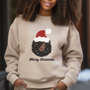 Könnte beinhalten: Ein beiger Pullover mit einem Aufdruck einer Frau mit Afrofrisur, die einen Weihnachtsmann-Hut trägt. Der Text "Merry Christmas" ist unter dem Aufdruck gedruckt.
