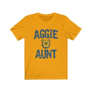May include: A gold t-shirt with a blue and white distressed graphic that says "Aggie Aunt" with a bulldog mascot in the middle.