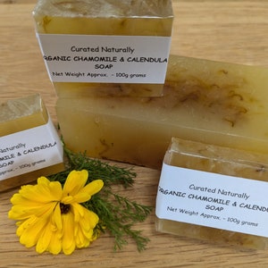 May include: Three bars of organic chamomile and calendula soap, each with a label that reads "Curated Naturally Organic Chamomile & Calendula Soap Net Weight Approx. - 100g grams". The soap is a translucent yellow colour with flecks of dried flowers.