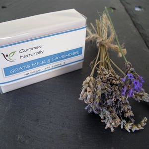 May include: A white bar of soap with a blue and white label that reads "Curated Naturally Goats Milk & Lavender Net Weight - 100 grams 3.5 oz". The soap is sitting on a black surface next to a bunch of dried lavender.