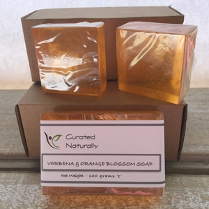 May include: Three bars of clear orange soap wrapped in plastic. The soap is labeled "Curated Naturally Verbena & Orange Blossom Soap Net Weight - 100 grams 'e'".