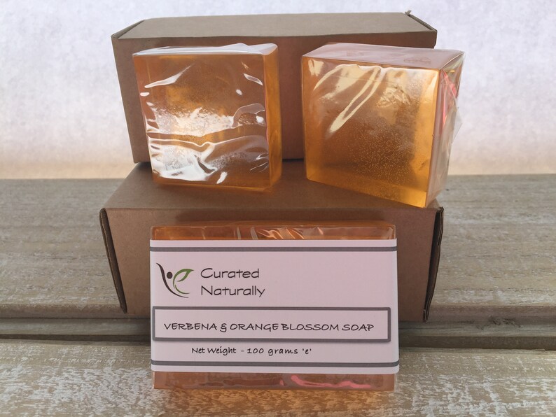 May include: Three bars of clear orange soap wrapped in plastic. The soap is labeled "Curated Naturally Verbena & Orange Blossom Soap Net Weight - 100 grams 'e'".