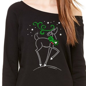 May include: Black off-the-shoulder sweatshirt with a green and silver sparkly reindeer design. The reindeer is holding a sprig of holly.