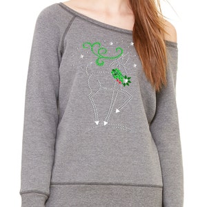 May include: A gray off-the-shoulder sweatshirt with a sparkly reindeer design featuring green holly and red berries. The reindeer is surrounded by white stars.