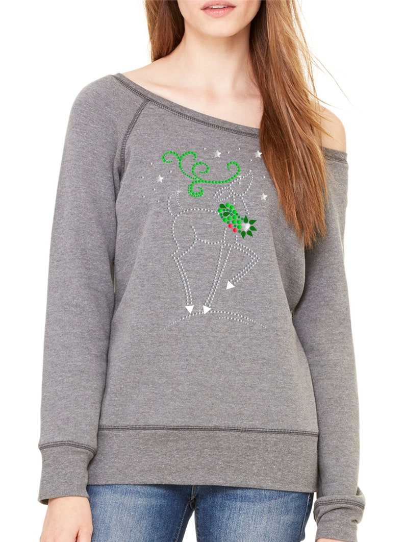 May include: A gray off-the-shoulder sweatshirt with a sparkly reindeer design featuring green holly and red berries. The reindeer is surrounded by white stars.