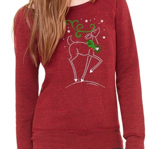 May include: Red long-sleeved sweatshirt with a sparkly reindeer design. The reindeer is wearing a green holly wreath and has a red heart on its nose. The design is surrounded by white stars.