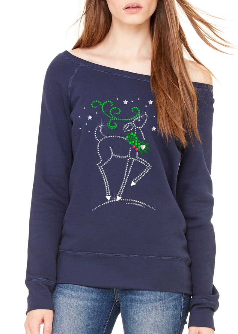 May include: A navy blue off-the-shoulder sweatshirt with a rhinestone reindeer design. The reindeer is holding a sprig of holly and is surrounded by silver glitter stars.