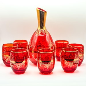 Könnte beinhalten: Ein rotes Glas-Dekanter- und Gläserset. Der Dekanter hat einen goldfarbenen Hals und Rand, und die Gläser haben goldfarbene Ränder. Das Set hat ein verziertes Goldmuster. Das Set enthält einen Dekanter und sechs Gläser.
