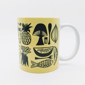 May include: A yellow ceramic mug with a black and white design of various fruits, vegetables, and other food items. The mug has a white handle.