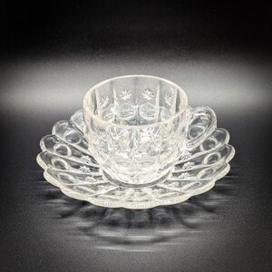May include: A clear glass teacup and saucer set with a cut glass design. The cup and saucer have a delicate, intricate pattern.