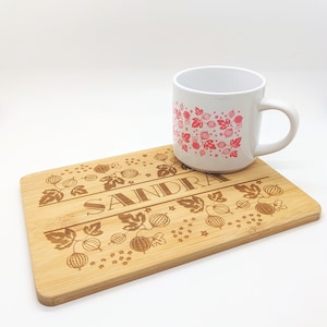 以下が含まれることがあります： 「SANDRA」という名前が刻まれた、パーソナライズされた竹製のまな板。まな板は葉とベリーのパターンで飾られています。ピンクのベリー柄の白いマグカップがまな板の上に置かれています。
