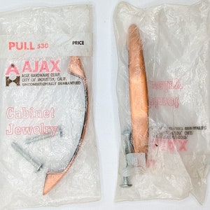 May include: Two copper cabinet pulls, individually packaged in plastic. The pulls are crescent moon shaped and have a satin finish. The packaging includes the text "PULL 530", "AJAX HARDWARE CORP.", "CITY OF INDUSTRY, CALIF.", "UNCONDITIONALLY GUARANTEED", and "Cabinet Jewelry".