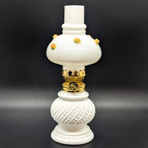 Puede incluir: Lámpara de aceite de vidrio blanco lechoso con un portamechas de metal dorado y una base blanca con un diseño texturizado. La lámpara tiene una pantalla de vidrio blanco con un borde festoneado y pequeñas flores naranjas y blancas.
