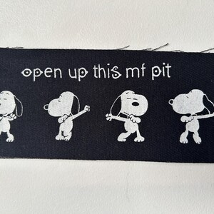 May include: Black fabric patch with white text that reads "open up this mf pit" and four white cartoon dogs in a dancing pose.