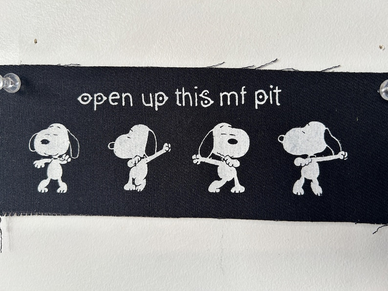 May include: Black fabric patch with white text that reads "open up this mf pit" and four white cartoon dogs in a dancing pose.