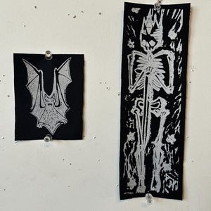 May include: Two black fabric patches pinned to a white wall. One patch features a white bat design, the other a white skeleton with a crown. The patches are rectangular and square.