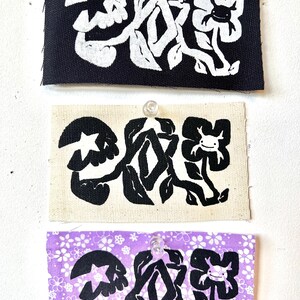 May include: Three fabric patches pinned to a white surface. Each patch features a black and white floral design with a star and a smiling flower. The patches are black, beige, and purple with a white floral pattern.
