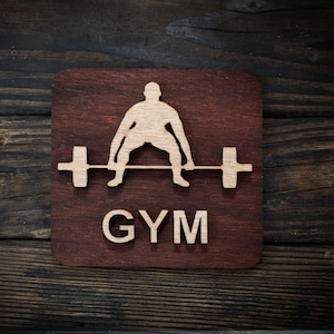 May include: A wooden sign with a brown background and a white silhouette of a person lifting a barbell. The word "GYM" is written in white letters below the silhouette.