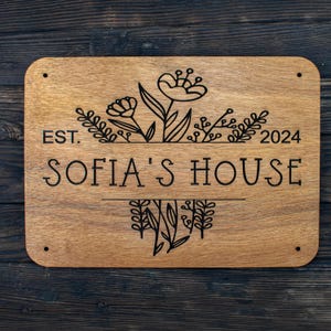 May include: A wooden sign with the text "EST. 2024 SOFIA'S HOUSE" and a floral design. The sign is brown and has a natural wood grain.