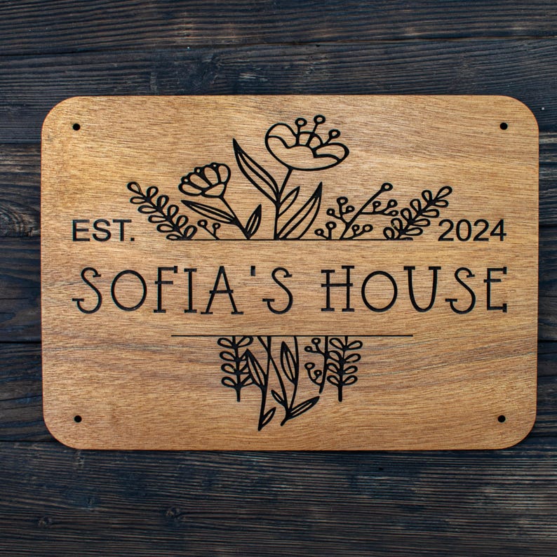 May include: A wooden sign with the text "EST. 2024 SOFIA'S HOUSE" and a floral design. The sign is brown and has a natural wood grain.