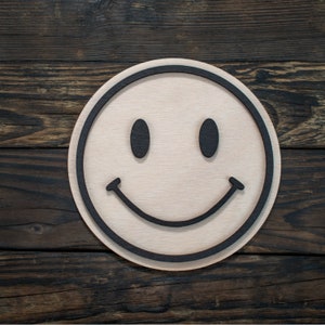 Peut inclure: Un visage souriant rond en bois avec un contour et des traits noirs. Le visage souriant est en bois de couleur claire avec des yeux noirs et une bouche incurvée. Le fond est une surface en bois brun foncé.