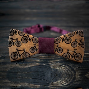 May include: A wooden bow tie with a brown background and black bicycle silhouettes. The bow tie has a burgundy adjustable strap.