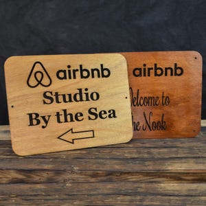 May include: Two wooden signs with the Airbnb logo and text. The sign on the left says "airbnb Studio By the Sea" with an arrow pointing left. The sign on the right says "airbnb Welcome to the Nook".