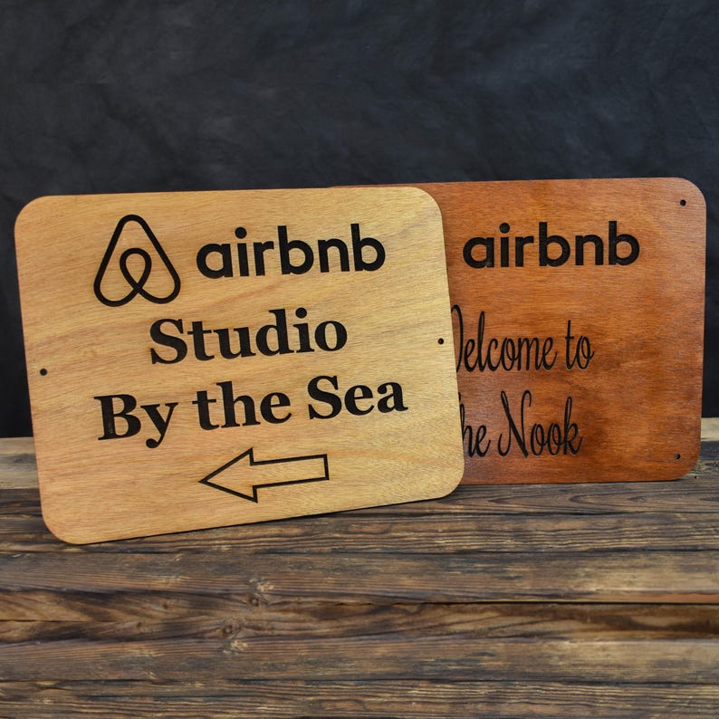 May include: Two wooden signs with the Airbnb logo and text. The sign on the left says "airbnb Studio By the Sea" with an arrow pointing left. The sign on the right says "airbnb Welcome to the Nook".