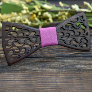 May include: A dark brown wooden bow tie with a wave-like cutout pattern. It features a bright pink fabric band in the centre. This wooden bow tie is a distinctive accessory for formal or casual attire.