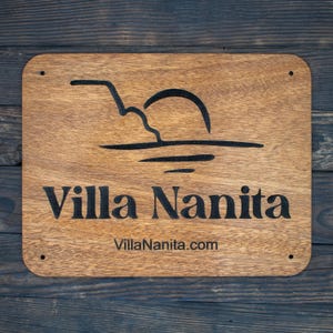 May include: A wooden sign with the text "Villa Nanita" and the website address "VillaNanita.com". The sign features a stylised image of a sunset over water.